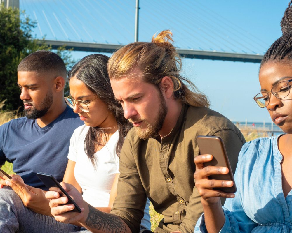 Focused multiracial friends using smartphones. Concentrated young male and female friends gathering together, sitting and using cell phones in park. Technology concept